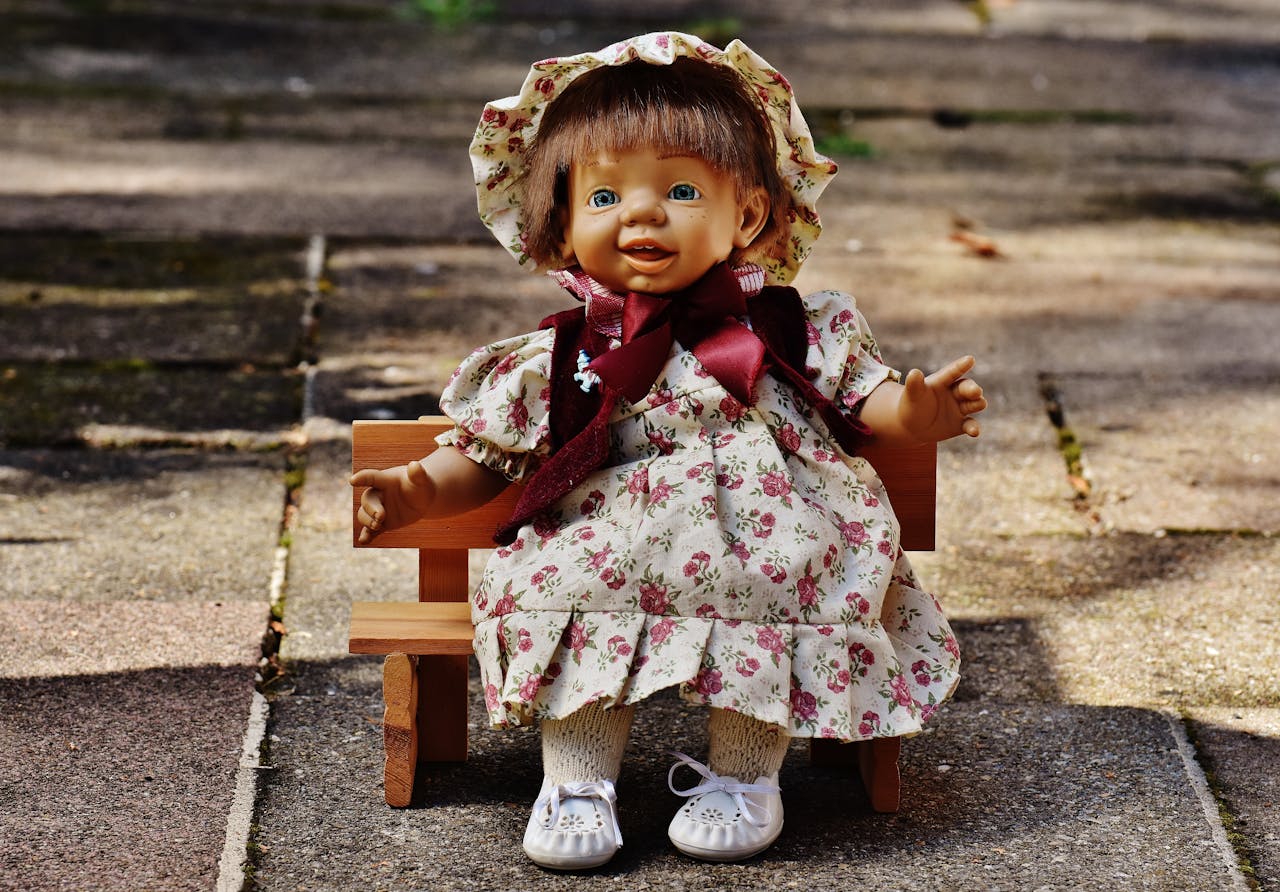Cute smiling doll wearing a floral dress and bonnet, sitting on a wooden bench outdoors.